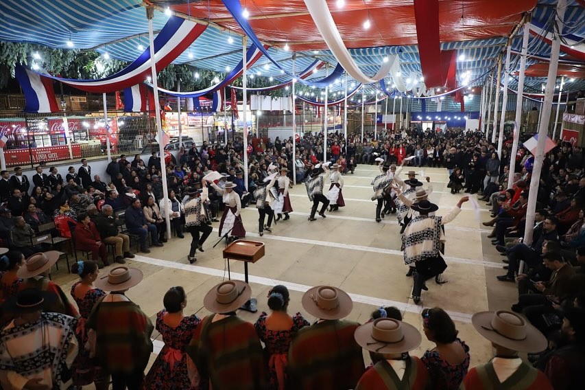 Bailarines de cueca vestidos con trajes de huaso y de china actuando frente a un público sentado bajo un toldo decorado con banderas chilenas.