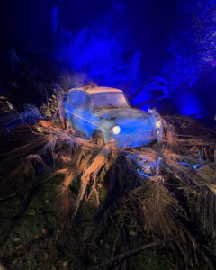 Un auto clásico de color azul rodeado de ramas secas y palmeras bajo una intensa iluminación azul, recreando una escena mágica en el Parque Quinta Normal.