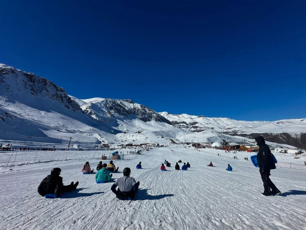 Grupo de personas sentadas en trineos deslizándose por una amplia pista de nieve en el Parque Farellones, bajo un cielo despejado de la Cordillera de los Andes.