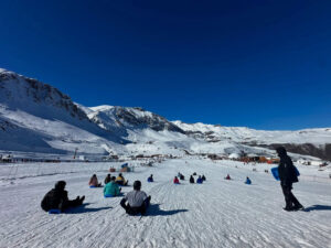Grupo de personas sentadas en trineos deslizándose por una amplia pista de nieve en el Parque Farellones, bajo un cielo despejado de la Cordillera de los Andes.