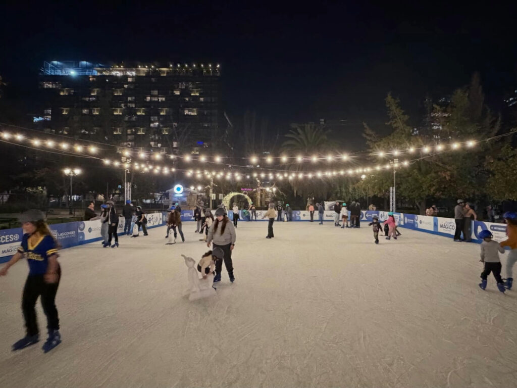 Niños y adultos patinando de noche en la pista de hielo al aire libre del Parque Araucano, decorada con guirnaldas de luces encendidas y edificios de fondo. 