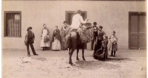 Fotografía histórica que captura el momento en que un vendedor a caballo entrega productos a una mujer y sus hijos frente a una casa de fachada continua. Esta escena refleja la importancia de los oficios chilenos antiguos en el abastecimiento y la vida urbana de los barrios de antaño.