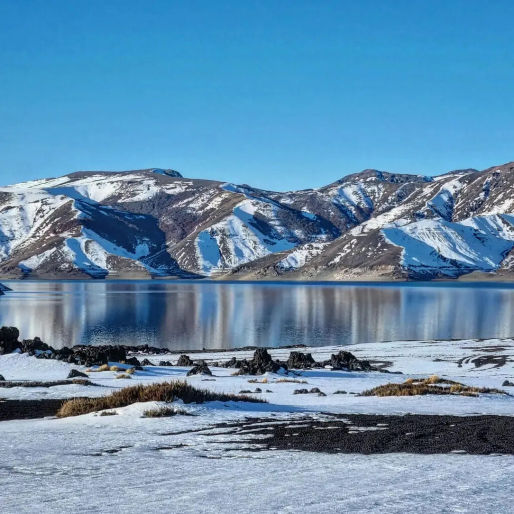 Aguas serenas de la Laguna del Laja reflejando las cumbres de las montañas nevadas vecinas, con rocas volcánicas oscuras y restos de nieve en la orilla.
