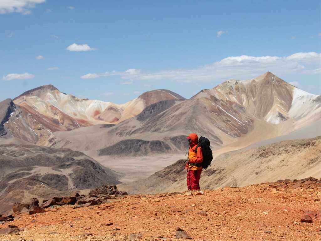 Persona con mochila de trekking y chaqueta naranja contempla las imponentes montañas de tonos rojizos, amarillos y ocres de Suriplaza, uno de los destinos naturales de Chile en el altiplano.