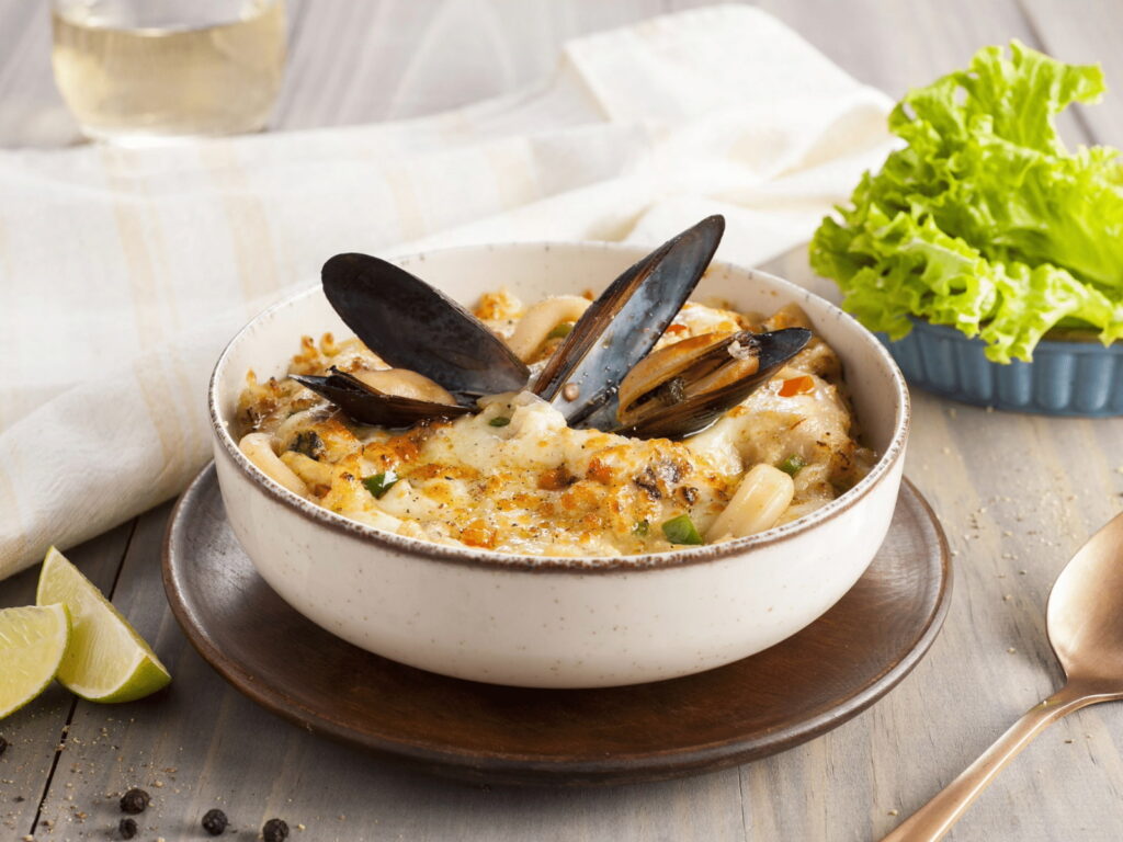 Un pocillo rústico de cerámica blanca con chupe de mariscos, decorado en el centro con tres choritos en su concha abierta. El plato está dispuesto sobre una mesa de madera clara junto a gajos de limón, pimienta negra en grano y una cuchara de cobre.