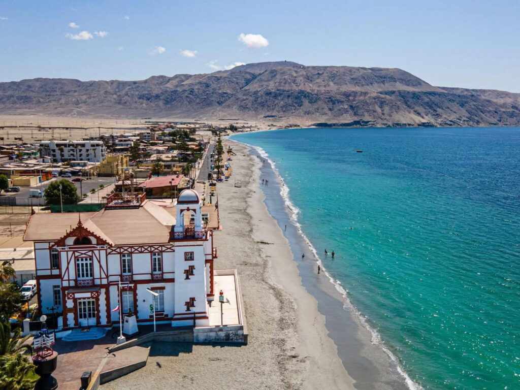 Toma aérea de la Capitanía de Puerto de Mejillones con su arquitectura roja y blanca, situada junto a una extensa playa de aguas turquesas y cerros desérticos. 