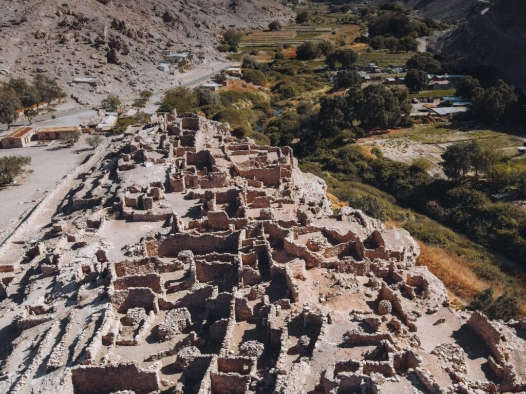 Vista aérea de los muros y pasajes de piedra del Pukará de Lasana, una antigua fortaleza prehispánica ubicada junto al verde oasis del valle en la Región de Antofagasta.