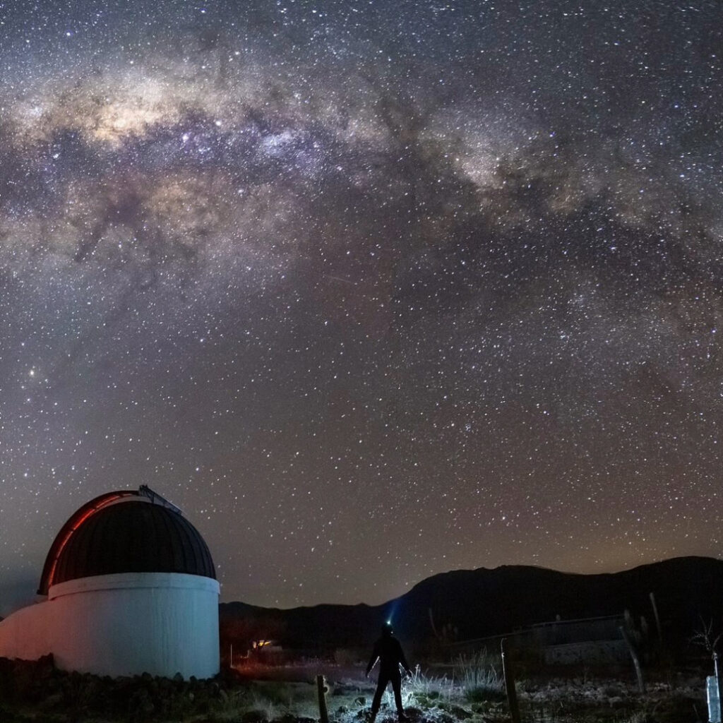 Una silueta humana con linterna frontal contempla la imponente Vía Láctea junto a una cúpula astronómica, viviendo una experiencia de astroturismo en la Región de Coquimbo.