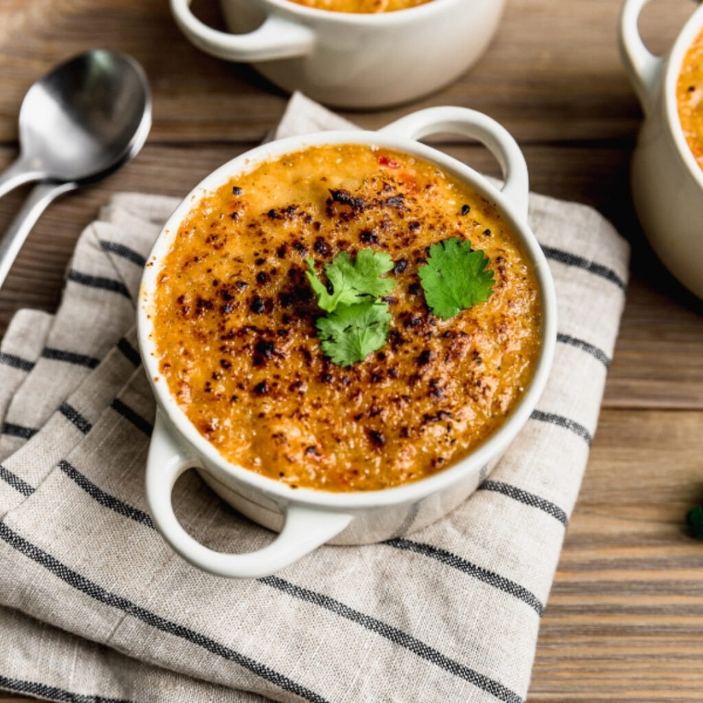 Un pocillo cerámico con asas que contiene una porción individual de chupe de mariscos bien gratinado, decorado con hojas de cilantro fresco en el centro. 