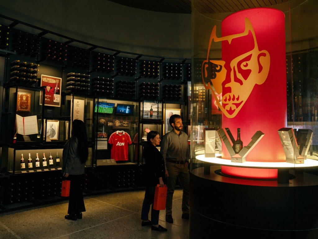Una pareja realiza un recorrido enoturístico guiado dentro de una moderna sala de exposición de la Viña Concha y Toro. A la derecha destaca una gran vitrina cilíndrica iluminada con la icónica figura del diablo en color rojo y oro para la línea Casillero del Diablo 