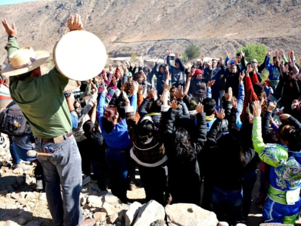 Un grupo de personas con los brazos alzados hacia el cielo participa en una ceremonia andina en Tierra Amarilla. El guía es un hombre que sostiene un tambor tradicional.