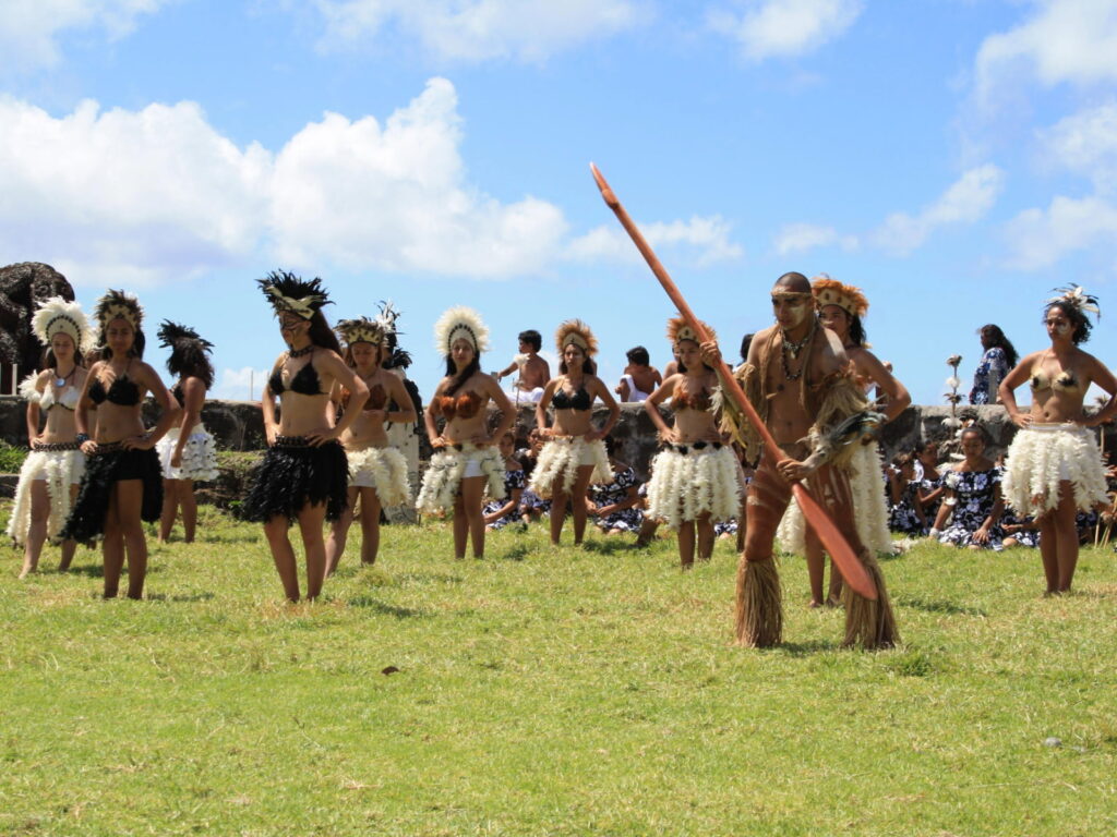 Un grupo de bailarines Rapa Nui con trajes de plumas y fibras naturales tradicionales realiza una danza ancestral sobre el pasto, bajo un cielo azul con nubes.