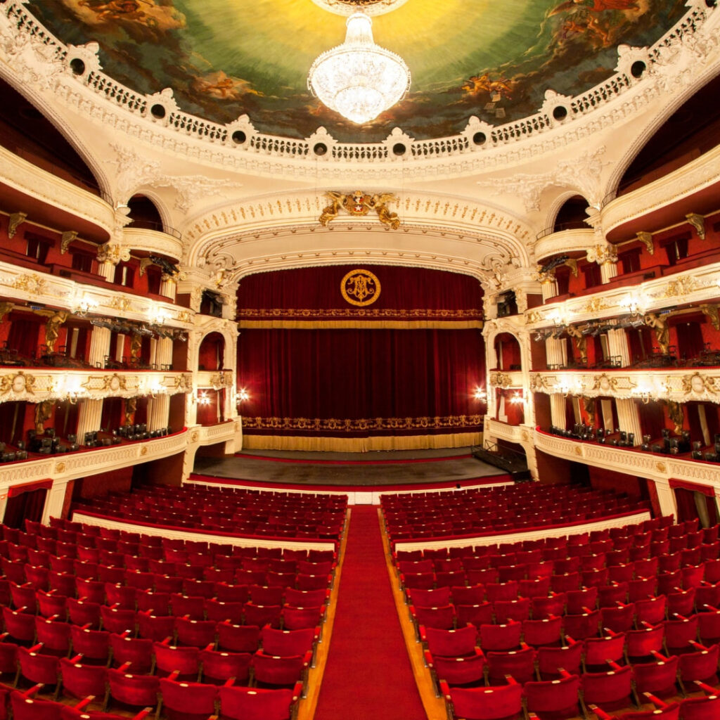 Vista panorámica en perspectiva central del opulento interior del Teatro Municipal de Santiago desde la platea. Se aprecian las corridas de butacas de terciopelo rojo perfectamente alineadas frente al imponente escenario con un telón rojo oscuro y detalles dorados. 