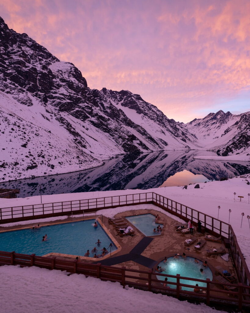 Turistas disfrutan de una piscina temperada y jacuzzis al aire libre en Ski Portillo, frente a la Laguna del Inca reflejando las montañas nevadas al atardecer.