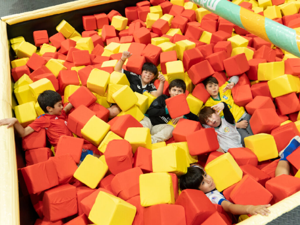 Un grupo de niños sonríe y saluda alegremente mientras se divierten sumergidos dentro de una gran piscina llena de cubos de espuma rojos y amarillos en Inspira Kids.