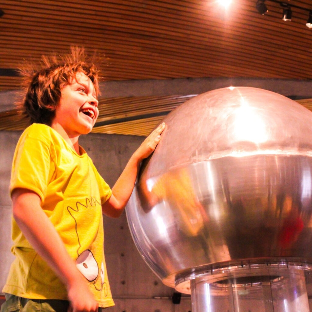 Un niño sonríe con asombro en el MIM mientras toca una esfera metálica conductora que genera electricidad estática, haciendo que su cabello se levante por completo.