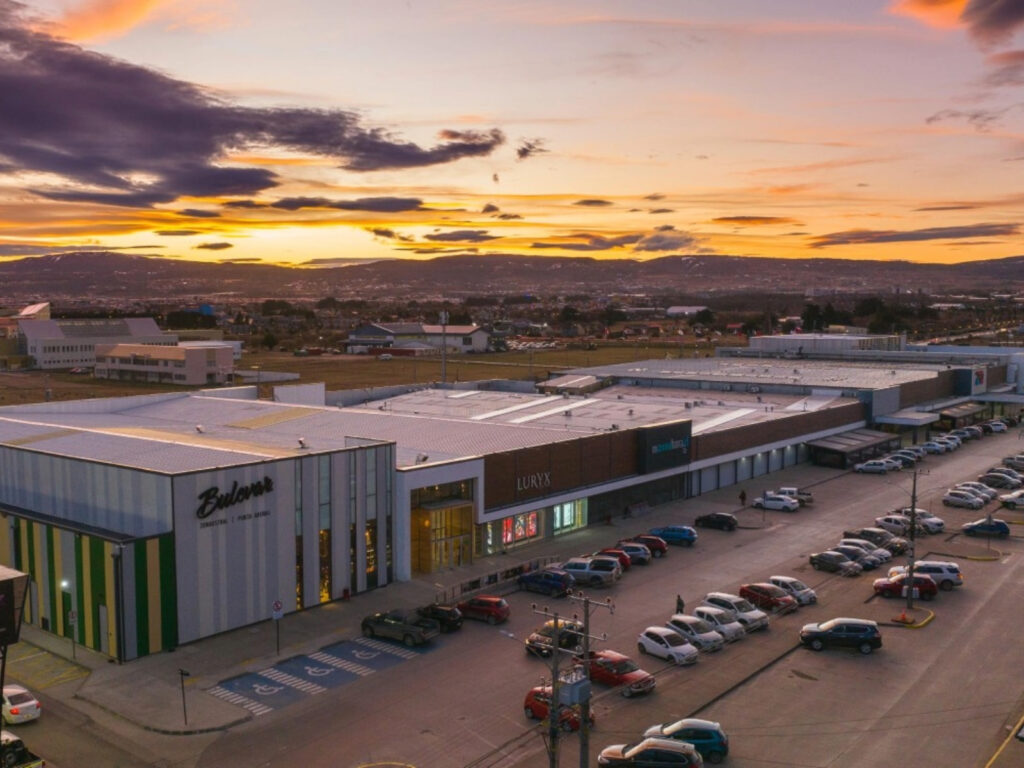Vista aérea del complejo comercial de la Zona Franca en Punta Arenas al atardecer. Se aprecia el estacionamiento con vehículos y de fondo los cerros iluminados por los tonos naranjos del cielo. 