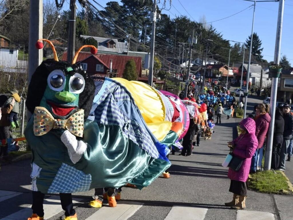 Una comparsa callejera avanza por una calle cuesta arriba durante la Puihua Cultural en Puyehue. En primer plano destaca una persona disfrazada con una gran cabeza de oruga verde con corbatín.
