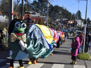Una comparsa callejera avanza por una calle cuesta arriba durante la Puihua Cultural en Puyehue. En primer plano destaca una persona disfrazada con una gran cabeza de oruga verde con corbatín.