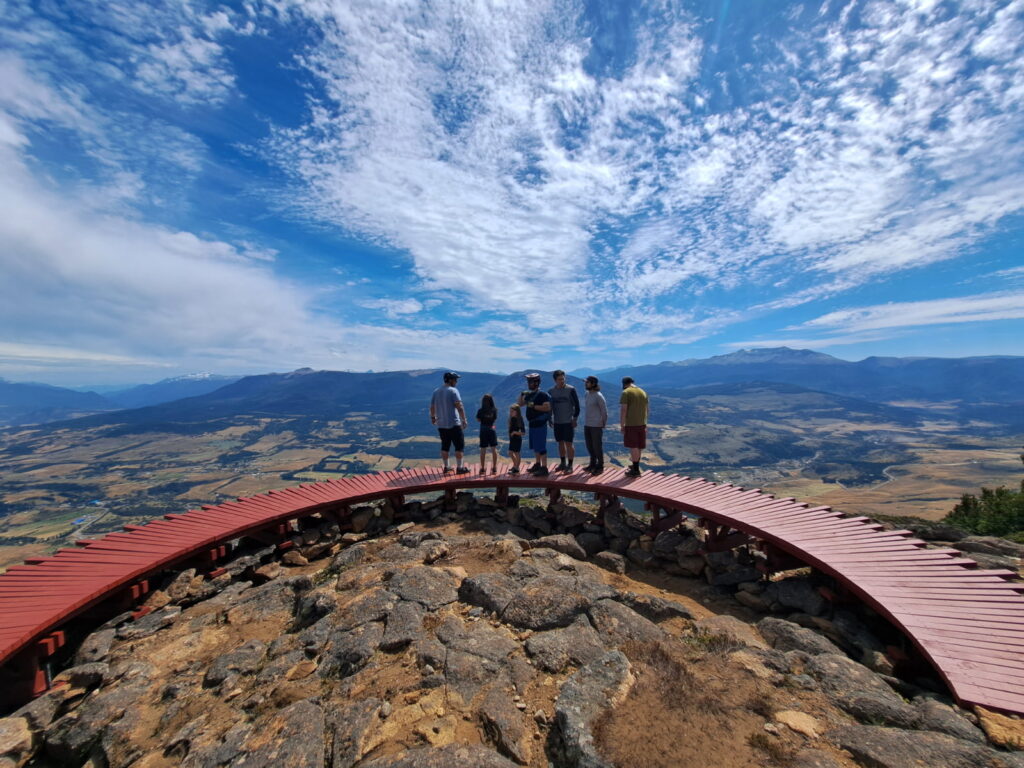  Un grupo de turistas de pie sobre una plataforma circular de madera en la cumbre del Parque El Cóndor, en Coyhaique. Al fondo, se aprecia una vista panorámica de un valle y montañas bajo un cielo azul con nubes ligeras.