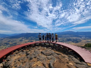 Un grupo de turistas de pie sobre una plataforma circular de madera en la cumbre del Parque El Cóndor, en Coyhaique. Al fondo, se aprecia una vista panorámica de un valle y montañas bajo un cielo azul con nubes ligeras.