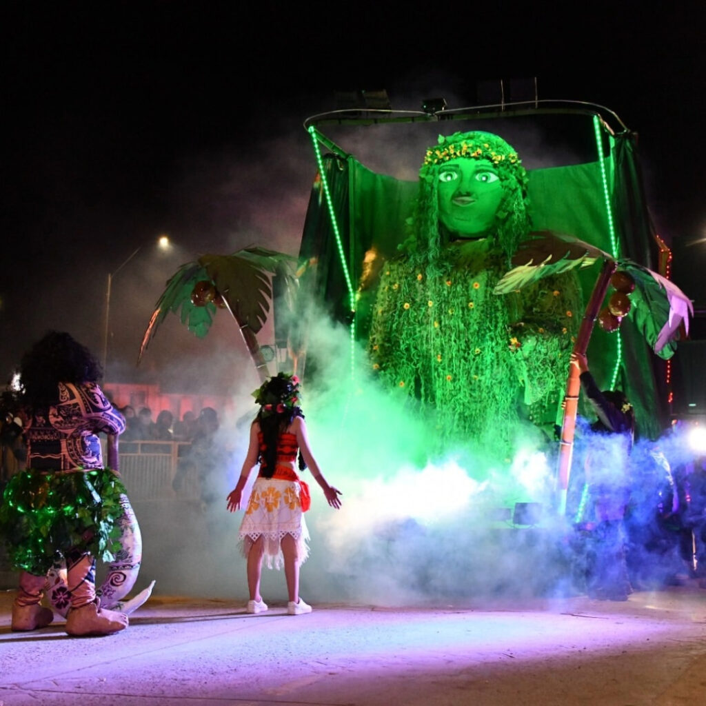 Carro alegórico iluminado de verde con una figura gigante en el Carnaval de Invierno de Punta Arenas de noche. En primer plano, personas con trajes tradicionales bailan entre humo blanco y luces. 