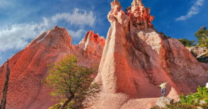 Una persona de pie sobre una roca con los brazos abiertos contempla las imponentes formaciones de Piedras Coloradas en Lonquimay, uno de los lugares secretos de Chile. El paisaje destaca por sus gigantescos cerros de roca volcánica teñida de un intenso color rojizo y un árbol nativo en primer plano bajo un cielo azul con nubes dispersas.