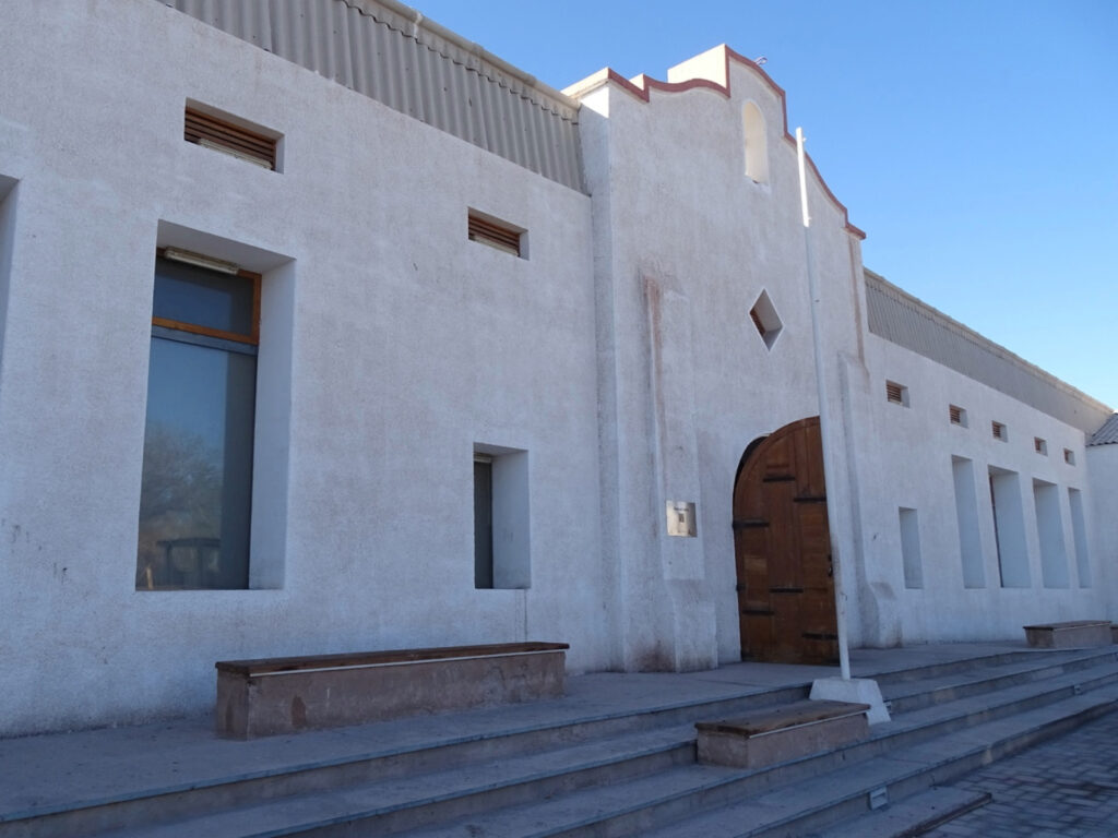Fachada de estilo colonial del Museo del Salitre en María Elena, Región de Antofagasta, con muros blancos, una gran puerta arqueada de madera y escaños de descanso en su entrada, ideal para complementar los panoramas bajo techo en Chile.