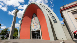 Fachada moderna de la Catedral de Chillán, con su característico arco de hormigón de color naranja, una cruz de concreto y un mural religioso sobre la puerta de entrada, frente a la Plaza de Armas.
