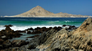 Costa rocosa con olas rompiendo en tonos turquesa y un cerro al fondo, paisaje típico del litoral desértico que recorre la Ruta Patrimonial Litoral de Atacama.