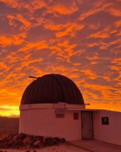 Silueta de la cúpula blanca de observatorio astronómico Cruz del Sur contra un cielo de atardecer intensamente encendido con nubes de color naranja y dorado brillante.