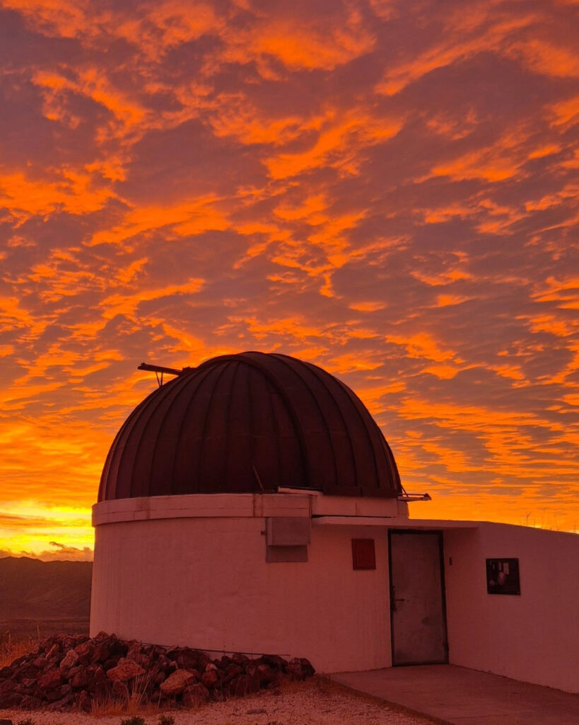 Silueta de la cúpula blanca de observatorio astronómico Cruz del Sur contra un cielo de atardecer intensamente encendido con nubes de color naranja y dorado brillante. 