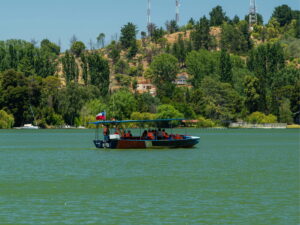 Una lancha a motor con turistas a bordo navega por las aguas verdes de la Laguna Avendaño en Quillón. Al fondo se observa una colina verde con árboles y antenas bajo un cielo despejado.