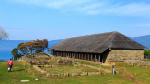 Antigua construcción de piedra y madera con techo de tejuelas junto a vestigios coloniales delimitados por muros bajos de piedra, uno de los sitios históricos de la Ruta Patrimonial Los Castillos del Fin del Mundo, con el estuario de Valdivia al fondo.