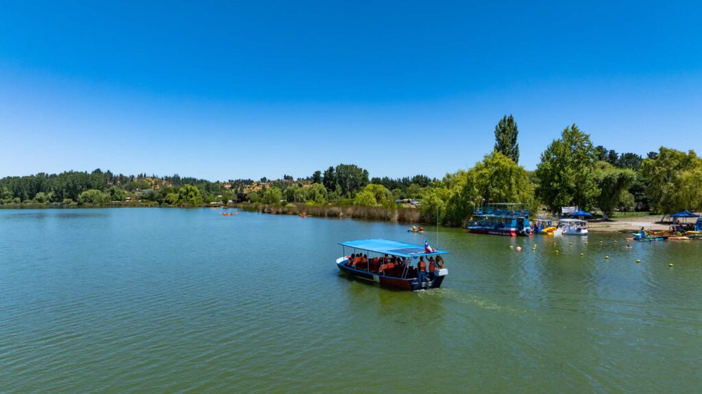 Una lancha a motor con turistas a bordo navega por las aguas verdes de la Laguna Avendaño en Quillón. Al fondo se observa una colina verde con árboles y antenas bajo un cielo despejado.