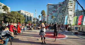 Artistas de circo actuando frente a un numeroso público de familias reunidas al aire libre en Viña del Mar, con edificios costeros, palmeras y banderines de colores de fondo, uno de los panoramas para el Día del Niño en Viña del Mar y Valparaíso.