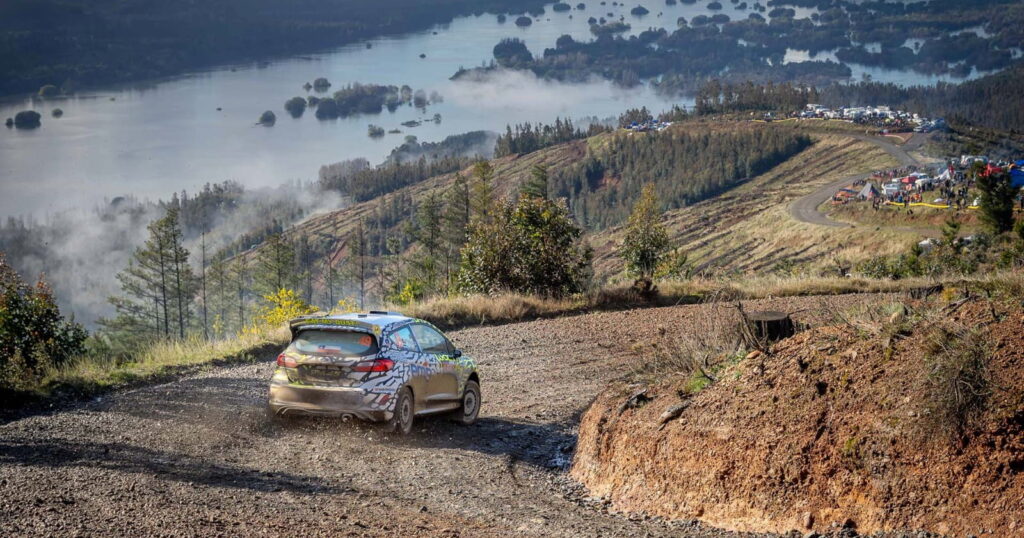 Auto de rally derrapando por un camino de ripio en las alturas, con un lago rodeado de bosques y espectadores observando desde la distancia, uno de los tramos del Rally Biobío 2026 en la Región del Biobío.