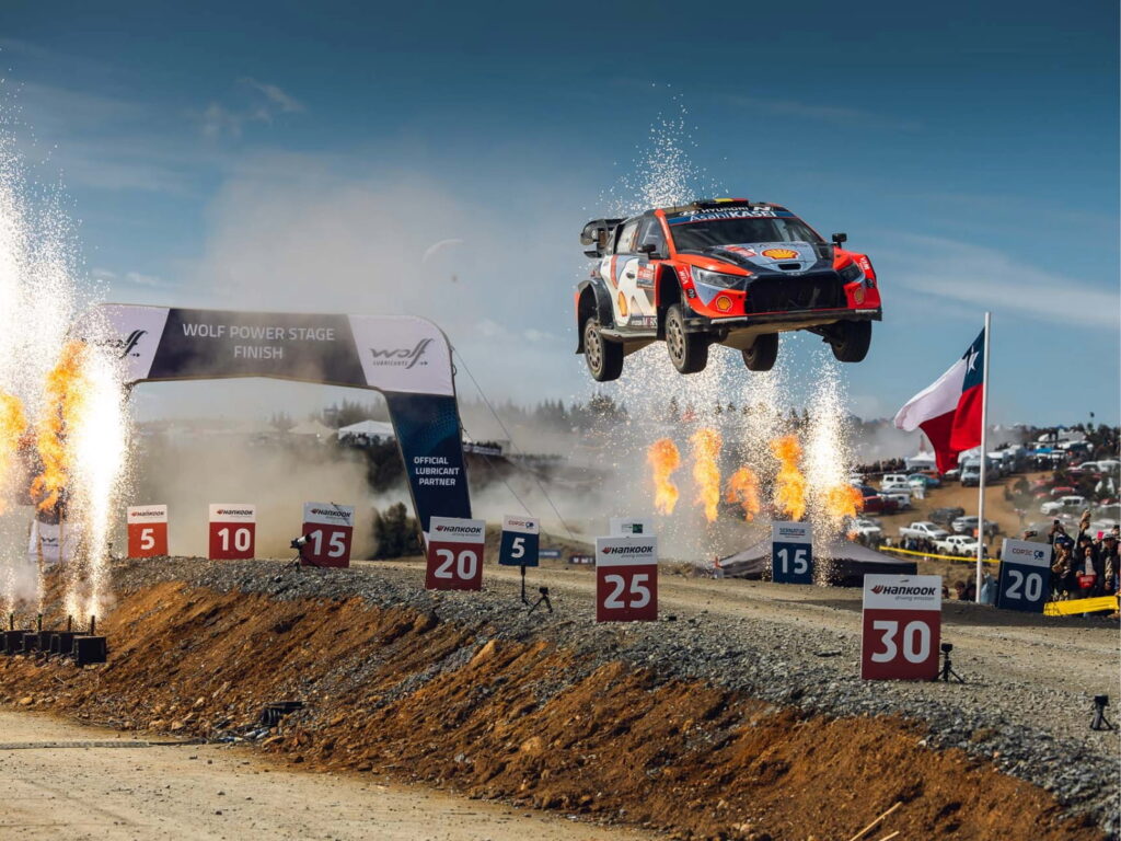 Auto de rally saltando en el aire entre llamas y polvo al cruzar el arco de meta del Wolf Power Stage, con espectadores y la bandera chilena de fondo.