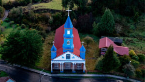 Vista aérea de la Iglesia de Tenaún, con su torre azul y techo rojo revestido en metal, rodeada de vegetación y una casa patrimonial, uno de los templos más pintorescos de la ruta por las Iglesias de Chiloé.