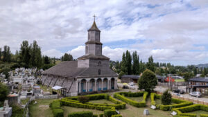 Vista aérea de la Iglesia de Nercón, de madera gris, rodeada de un cementerio con jardines y setos recortados, uno de los templos patrimoniales cercanos a Castro.
