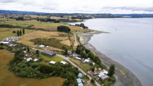 Vista aérea de la Iglesia de Vilupulli junto a la costa, con su esbelta torre, rodeada de campos y casas dispersas frente a las aguas tranquilas del mar interior de Chiloé.