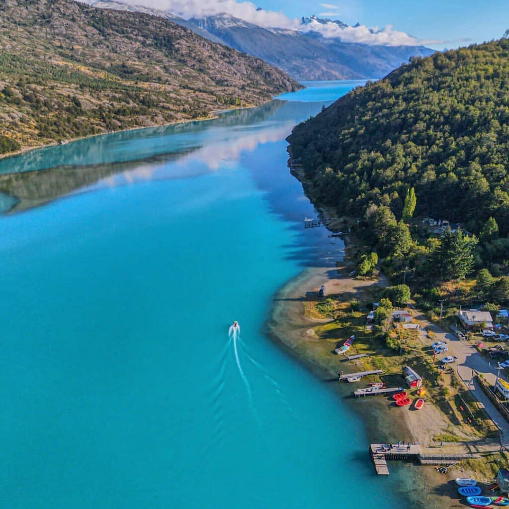 Vista aérea de las aguas turquesas del lago Bertrand, en la Región de Aysén, con un bote surcando la orilla y el pequeño poblado de Puerto Bertrand junto a la costa.