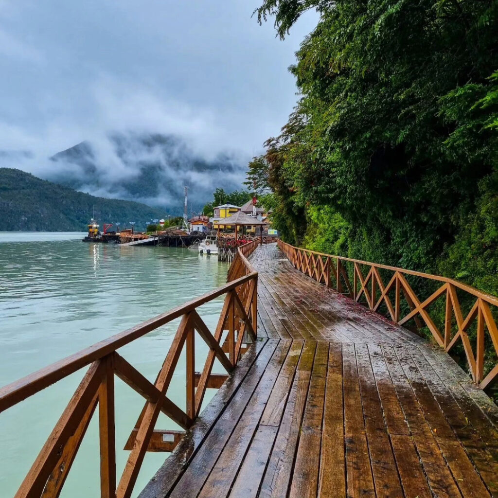 Pasarela de madera con baranda que bordea el fiordo de Caleta Tortel, con embarcaciones ancladas, casas de colores y montañas envueltas en niebla al fondo.
