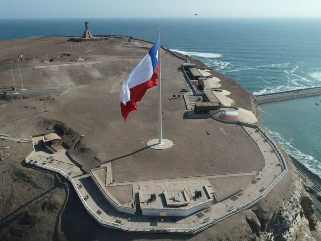 Vista aérea del Museo de Armas del Morro de Arica, con la gran bandera chilena ondeando junto al mirador, cañones históricos, terrazas fortificadas y el océano Pacífico de fondo.