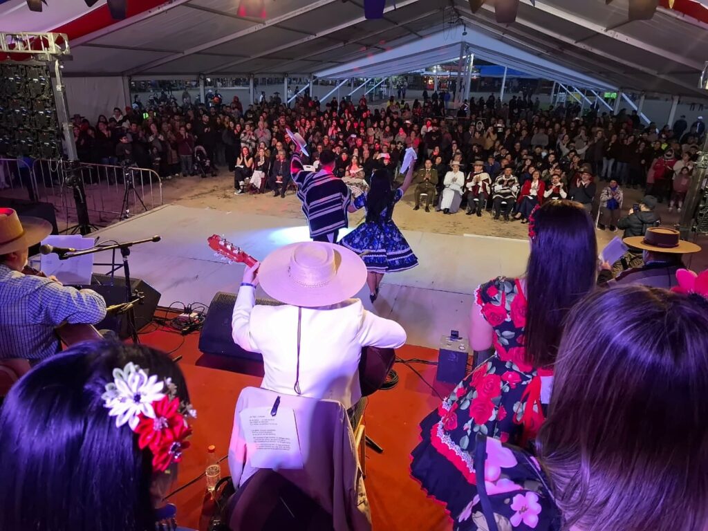 Carpa de cueca: pareja bailando en el escenario de una fonda techada, rodeada de público que aplaude durante las Fiestas Patrias. 