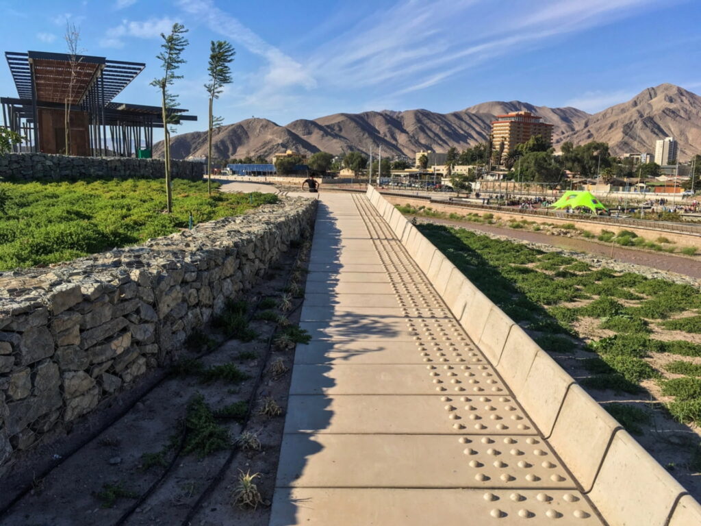 Sendero de concreto junto al cauce del río Copiapó en el Parque Kaukari, con muros de gaviones de piedra, jardines de vegetación desértica y cerros áridos con la ciudad de Copiapó al fondo, uno de los destinos para escapadas de fin de semana sin auto.