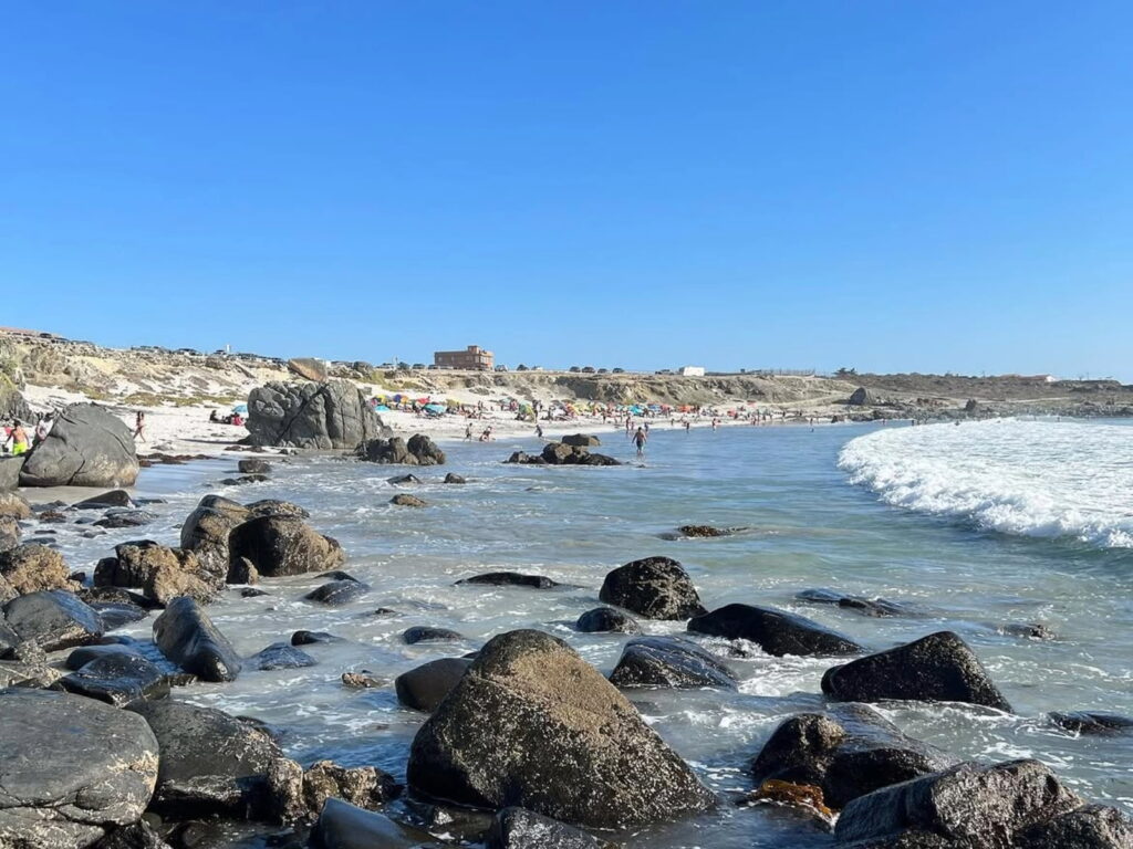 Playa rocosa de Punta de Choros con grandes piedras oscuras en primer plano, olas rompiendo en la orilla y bañistas disfrutando de la arena blanca junto a la costa, en la Región de Coquimbo.