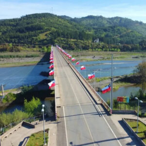 Vista aérea del Puente Paula en Hualañé, completamente engalanado con banderas chilenas a ambos lados, sobre un río rodeado de cerros boscosos.