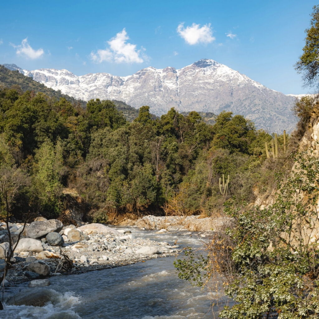 Un río de aguas claras corre entre piedras y vegetación nativa, con la cordillera nevada de fondo, en un paisaje típico de la zona central chilena durante la primavera en Chile.
