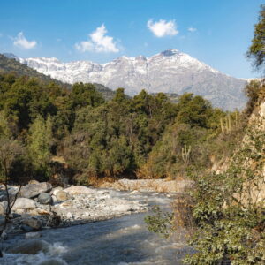 Un río de aguas claras corre entre piedras y vegetación nativa, con la cordillera nevada de fondo, en un paisaje típico de la zona central chilena durante la primavera en Chile.